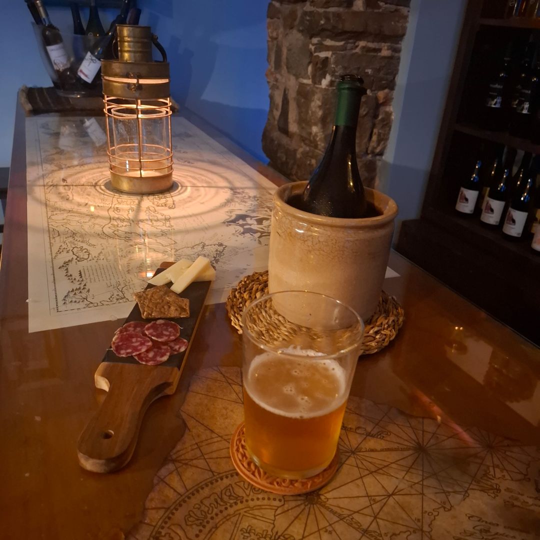 Aperitivo sul bancone in vetro: una lanterna d'ottone proietta cerchi di luce su una carta nautica accanto a salame, formaggio, una birra e una bottiglia in glacette.