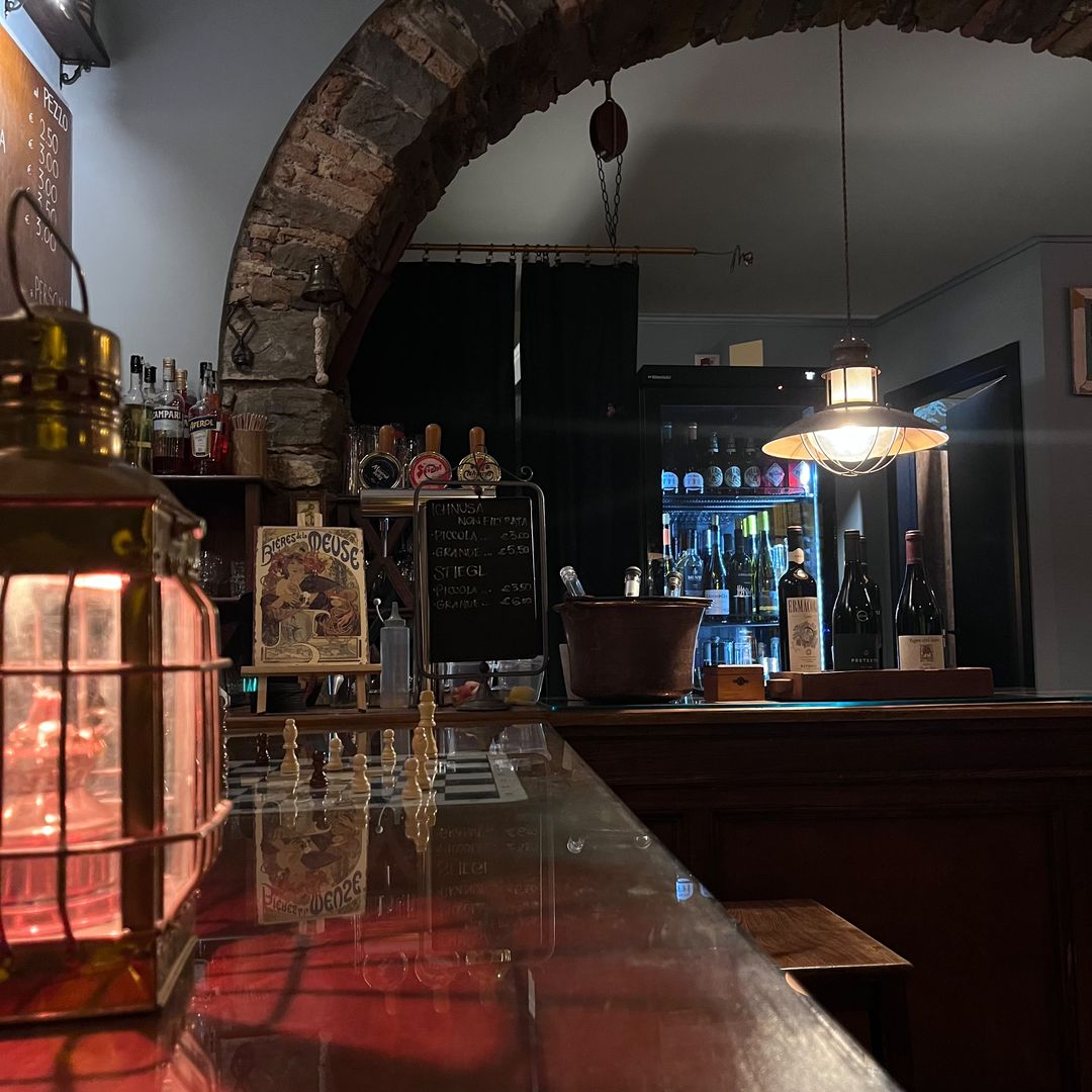 Lanterna d'ottone accesa sul bancone in vetro con pezzi degli scacchi; sullo sfondo l'arco in pietra, le spine della birra e una stampa liberty.
