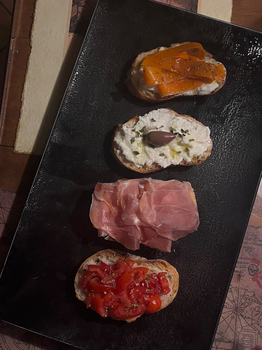 Quattro crostini su un piatto nero: carote marinate e stracciatella, stracciatella con oliva, prosciutto crudo, pomodorini.