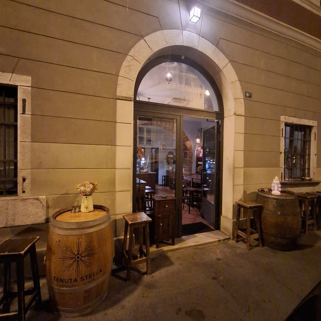 Facciata di Trebisonda di notte in Via dell'Annunziata 6C: portale ad arco aperto, botti e sgabelli in legno sul marciapiede.