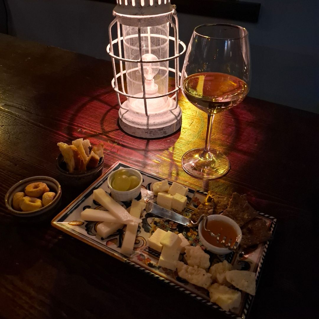 Calice di vino bianco accanto a una lanterna accesa e a un piatto in ceramica dipinta con formaggi, miele, uva e cracker.
