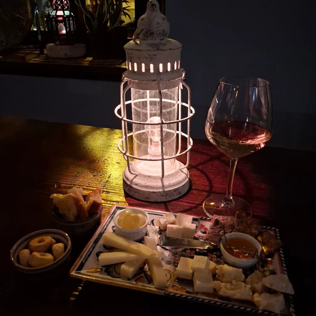 Calice di vino rosato, lanterna accesa e piatto di formaggi con miele, uva e cracker su un tavolo scuro.