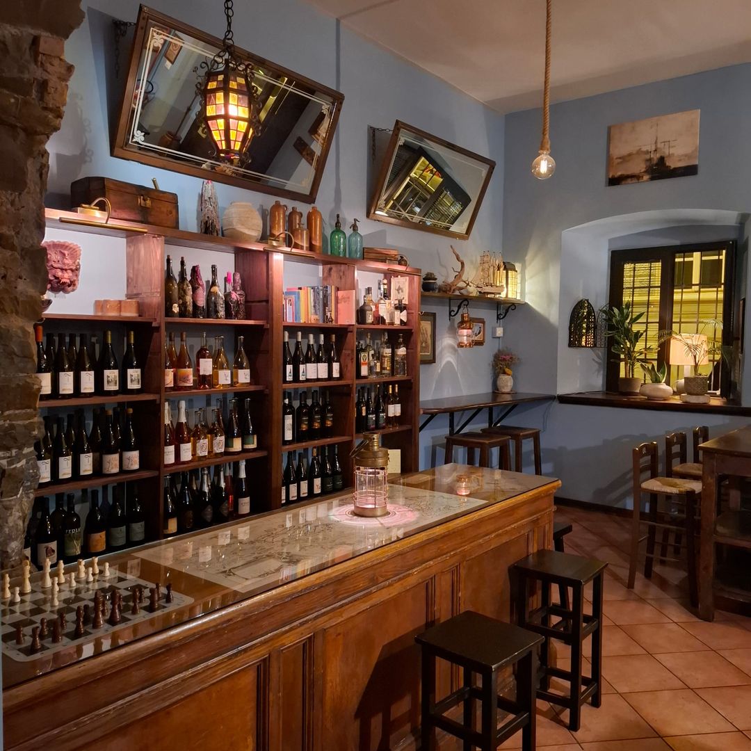 La parete dei vini di Trebisonda: grande scaffale in legno pieno di bottiglie, specchi inclinati e bancone in vetro con una carta nautica, una lanterna e una scacchiera.