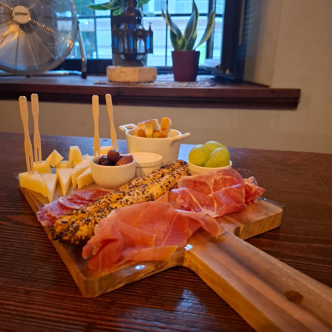 Tagliere su pala di legno con prosciutto crudo, salame, cubetti di formaggio, olive, grissini ai semi e uva bianca.